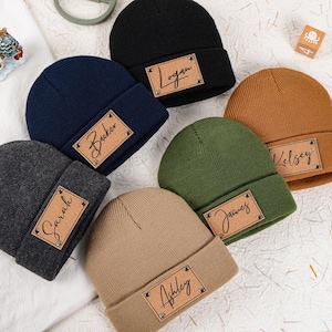 Puede incluir: Una colección de gorros personalizados en varios colores, incluyendo negro, azul marino, gris, verde oliva y beige. Cada gorro presenta un parche rectangular de cuero con un nombre en escritura cursiva. Los gorros están colocados sobre una superficie blanca.
