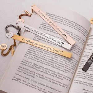 May include: A collection of gold and black metal bookmarks with inspirational quotes engraved on them. The bookmarks are displayed on a book with a white background.
