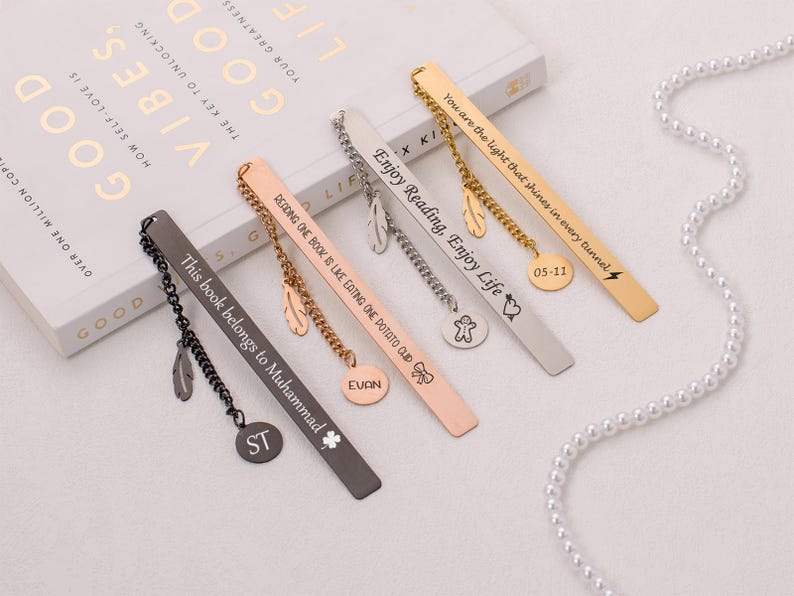 May include: A set of six personalized metal bookmarks with different engraved messages and charms. The bookmarks are black, silver, gold, and rose gold. The charms include a feather, a heart, a lightning bolt, a star, and a small figure.