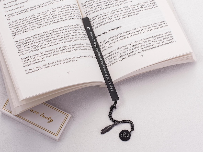 May include: A black metal bookmark with a feather charm and a double heart charm. The bookmark has an inscription that reads "People who love learning are always a joy to be along!"
