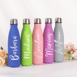 Könnte beinhalten: Fünf farbenfrohe, isolierte Wasserflaschen mit silbernen Deckeln. Jede Flasche hat eine andere Farbe: blau, grün, rosa, lila und grau. Jede Flasche hat einen Namen in weißer Schrift: Barbara, Madison, Cecilia, Marcus und William. Blumen sind im Vordergrund.