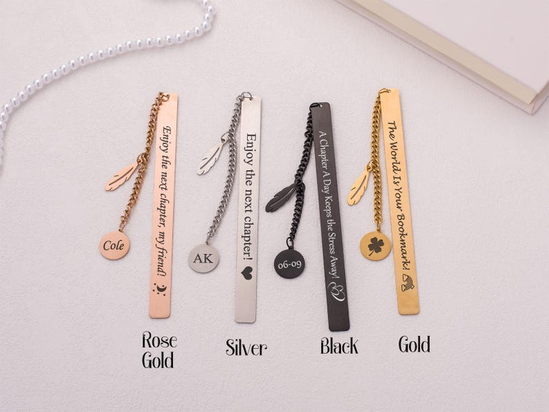 May include: Four metal bookmarks with different colored finishes: rose gold, silver, black, and gold. Each bookmark has a chain with a charm and a personalized inscription. The inscriptions are: "Enjoy the next chapter, my friend!" "Enjoy the next chapter!" "A Chapter A Day Keeps the Stress Away!" and "The World Is Your Bookmark!"