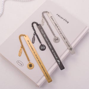 May include: Three gold and silver bookmarks with different engraved sayings. The gold bookmark has a clover charm and a feather charm. The silver bookmarks have feather charms. The sayings on the bookmarks are "I'm not ignoring you, I'm just reading something better", "Enjoy the next chapter, my friend!" and "Exponame is simply the name to go on a wilder - Jim Hills".