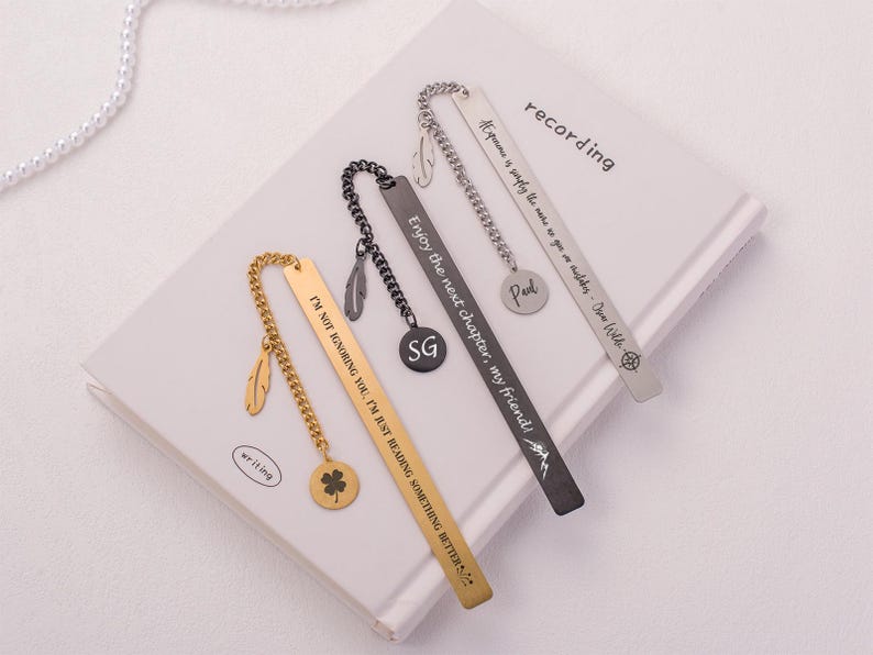 May include: Three gold and silver bookmarks with different engraved sayings. The gold bookmark has a clover charm and a feather charm. The silver bookmarks have feather charms. The sayings on the bookmarks are "I'm not ignoring you, I'm just reading something better", "Enjoy the next chapter, my friend!" and "Exponame is simply the name to go on a wilder - Jim Hills".