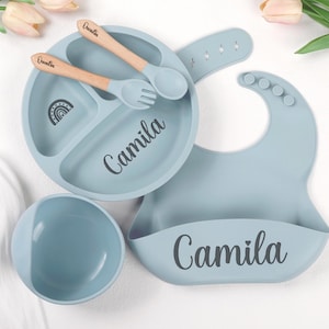 Könnte beinhalten: Ein staubblaues Silikon-Esslernset bestehend aus einem unterteilten Teller mit Regenbogen-Symbol und dem Namen Camila, einer passenden Schale, einem Lätzchen mit dem Namen Camila sowie einem Holzlöffel und einer Gabel mit dem Markennamen Quentin.