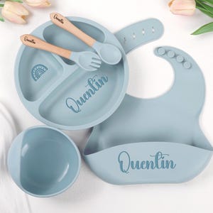 Op de afbeelding: Een babyvoedingsset in zachtblauwe kleur, inclusief een verdeelde bord, kom, slab en keukengerei met houten handvatten. Het bord heeft een regenboogontwerp en de naam "Quentin". De slab heeft een zak.