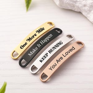 May include: Four rectangular metal tags with rounded ends and holes on each side. The gold tag reads One More Mile, the black tag reads Make It Happen, the silver tag reads KEEP RUNNING, and the rose gold tag reads You Are Loved in black text.