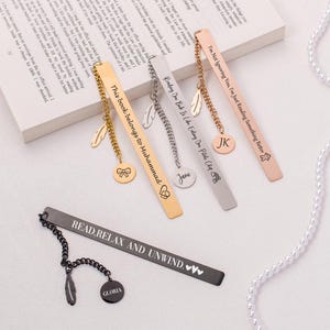 May include: Five gold, silver, and black metal bookmarks with different engraved messages and charms. The bookmarks have a feather charm and a round charm with a name or message. The bookmarks are on a white surface with a string of white pearls.
