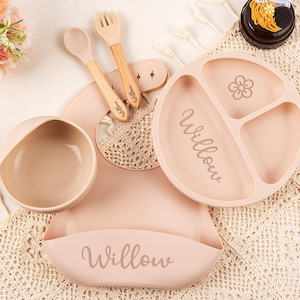 Peut inclure: Un ensemble d'alimentation pour bébé rose clair, comprenant une assiette divisée, un bol, un bavoir, une cuillère et une fourchette. L'assiette présente un motif floral et le nom "Willow". La cuillère et la fourchette ont des manches en bois.