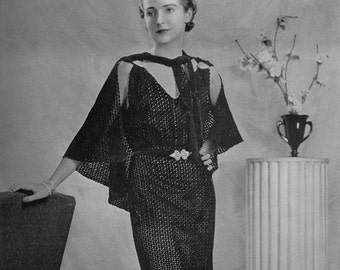 May include: A black crochet dress with a cape-like top and a belt. The dress is fitted and has a long skirt. The model is wearing the dress and standing in front of a white column.