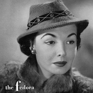 May include: A woman wearing a gray crocheted fedora with a black and white ribbon and decorative accents. The fedora has a wide brim and a slightly pointed crown. The text "the fedora" is printed below the woman's head.