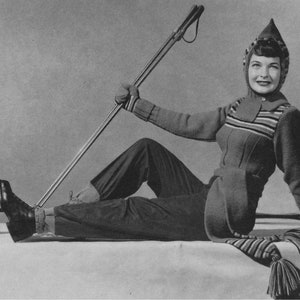 Può includere: Una donna indossa una tuta da sci a righe con un cappello e una sciarpa abbinati e posa con un bastone da sci. La tuta è di colore grigio scuro con strisce bianche e rosse. Il cappello è a punta con un pon pon bianco. La sciarpa è lunga e ha un fiocco all'estremità.