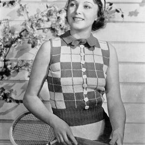 May include: A woman wearing a white and black checkered sleeveless top with a white collar and buttons. She is holding a tennis racket and is standing in front of a white wooden wall.