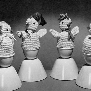 May include: Four crocheted chicks in egg cups. Each chick is wearing a different hat and scarf. The chicks are white with black eyes and beaks.
