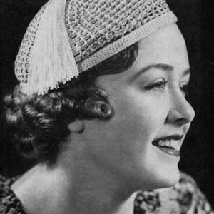 May include: A vintage-style crocheted hat with a textured pattern and a decorative tassel. The hat is light-colored and appears to be made of a soft yarn. The hat is worn by a person with curly hair.