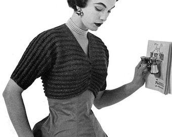 May include: A vintage black and white photograph of a woman wearing a dark, ribbed, short-sleeved sweater over a dress. She is wearing a pearl necklace, earrings, and a bracelet. She is holding a playbill and binoculars.
