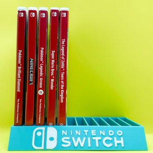 May include: A turquoise blue Nintendo Switch game stand with the text "NINTENDO SWITCH" in white. The stand holds six video game cartridges, including "The Legend of Zelda: Tears of the Kingdom", "Super Mario Bros. Wonder", "Pokémon Legends: Arceus", "Minecraft", and two Pokémon games.