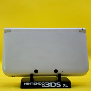 May include: White Nintendo 3DS XL handheld gaming console on a black stand with the text "Nintendo 3DS XL" printed on the front.