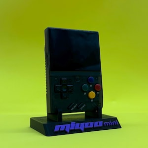 May include: A black handheld gaming console with a screen, directional pad, and colored buttons. The console is on a black stand with the text "miyoo mini" in purple. The background is a solid yellow.
