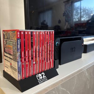 Puede incluir: Un soporte negro para juegos de Nintendo Switch con el logotipo de Nintendo Switch, que contiene varias carcasas de juegos rojas. Los juegos incluyen Pokémon y The Legend of Zelda. También se ve una consola Nintendo Switch negra.