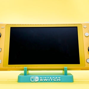 Może przedstawiać: Żółta konsola Nintendo Switch z czarnym ekranem stoi na niebieskim stojaku z napisem "Nintendo Switch".