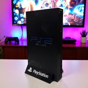 May include: Black PlayStation 2 console with the PS2 logo in blue. The console is displayed on a white surface with a black stand that has the PlayStation logo. The background includes a TV and a controller.