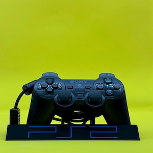 Op de afbeelding: Een zwarte PlayStation 2-controller met een zwarte standaard waarop "PS2" in blauw staat. De controller heeft een grijze analoge stick en knoppen.