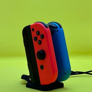 May include: Two Nintendo Switch Joy-Con controllers, one red and one blue, are plugged into a charging dock. The dock is black and has a cable attached to it.