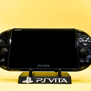 May include: A black PlayStation Vita handheld gaming console with a black stand. The console has a black screen and buttons. The stand has the PlayStation Vita logo.