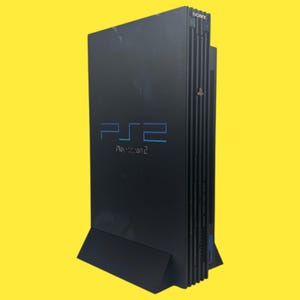 Peut inclure: Console PlayStation 2 noire avec le logo PS2 en bleu. La console est debout sur un support noir. Le logo Sony est visible dans le coin supérieur droit de la console.