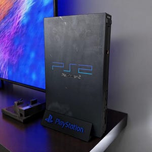 Peut inclure: Une console PlayStation 2 noire avec le logo "PS2" en bleu. La console est présentée sur une surface sombre avec un support logo PlayStation. Un appareil de jeu classique.