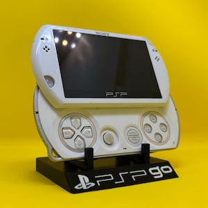 Puede incluir: Una consola de juegos portátil Sony PSP Go blanca sobre un soporte negro. La consola tiene una pantalla negra y botones de control. El soporte tiene el texto "PSP go" en blanco. El fondo es amarillo.