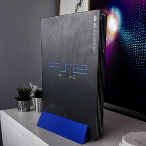 Peut inclure: Une console PlayStation 2 noire avec le logo et le texte "PlayStation 2" en bleu. La console est soutenue par un support bleu. La console est posée sur une surface blanche avec de petites plantes en pot.