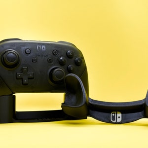 May include: A black Nintendo Switch Pro Controller stand with a black base and two black arms that hold the controller in place. The stand has the Nintendo Switch logo on the front.
