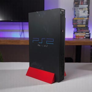 May include: A black PlayStation 2 console with the text "PS2" in blue and "PlayStation 2" below it. The console is on a red stand. The Sony logo is visible on the top right corner.