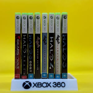 May include: A selection of Xbox 360 game cases, featuring titles such as "Gears of War," "Halo," and "Batman: Arkham." The games are presented in a white stand with the Xbox 360 logo. The cases have green tops and diverse game artwork.