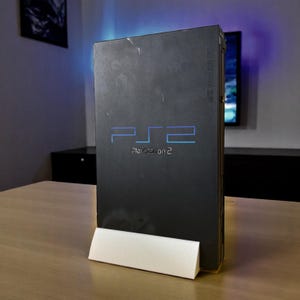 May include: A black PlayStation 2 console with the text "PS2" in blue and "PlayStation 2" below. The console is displayed on a white stand on a wooden table. The background includes a television and wall art.
