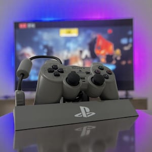 以下が含まれることがあります： PlayStation 2のグレーのコントローラーと、PlayStationのロゴが入ったスタンド。コントローラーには黒いアナログスティックとボタンがあります。背景にはビデオゲームを表示するテレビがあります。