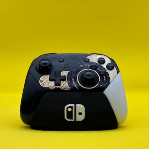 May include: A black and gold Nintendo Switch controller with a unique design featuring a circular pattern. The controller has a black and white Nintendo logo on the front.