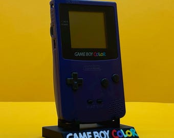 May include: A purple Game Boy Color handheld video game console on a black stand with the words "GAME BOY COLOR" in colorful letters.
