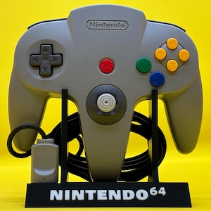 Puede incluir: Un mando gris de Nintendo 64 con botones de colores, expuesto sobre un soporte negro. El soporte tiene las palabras "NINTENDO 64" en blanco. El mando tiene un botón de inicio rojo, una cruceta y un joystick.