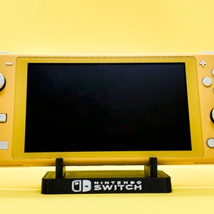 Może przedstawiać: Żółta przenośna konsola do gier Nintendo Switch z czarnym ekranem, stojąca na czarnym stojaku z nadrukiem "Nintendo Switch".
