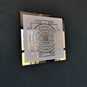 Puede incluir: Un cuadrado blanco enmarcado en oro con un dise&ntilde;o geom&eacute;trico en el centro. El dise&ntilde;o es un Sri Yantra, un s&iacute;mbolo sagrado en el hinduismo.