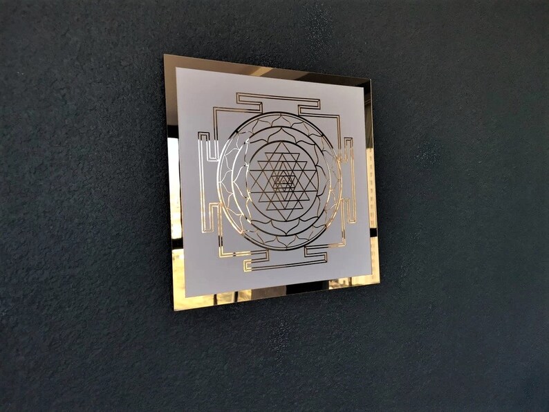 Puede incluir: Un cuadrado blanco enmarcado en oro con un dise&ntilde;o geom&eacute;trico en el centro. El dise&ntilde;o es un Sri Yantra, un s&iacute;mbolo sagrado en el hinduismo.