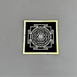 Puede incluir: Un cuadrado negro con un borde dorado, que presenta un s&iacute;mbolo Sri Yantra blanco. El s&iacute;mbolo es un dise&ntilde;o geom&eacute;trico complejo con un punto central y m&uacute;ltiples tri&aacute;ngulos.