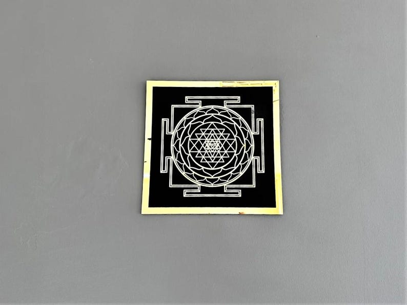 Puede incluir: Un cuadrado negro con un borde dorado, que presenta un s&iacute;mbolo Sri Yantra blanco. El s&iacute;mbolo es un dise&ntilde;o geom&eacute;trico complejo con un punto central y m&uacute;ltiples tri&aacute;ngulos.