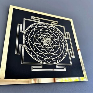Puede incluir: Una pieza de arte de pared cuadrada con un marco dorado, fondo negro y un dise&ntilde;o geom&eacute;trico blanco. El dise&ntilde;o presenta un mandala central con un s&iacute;mbolo Sri Yantra.