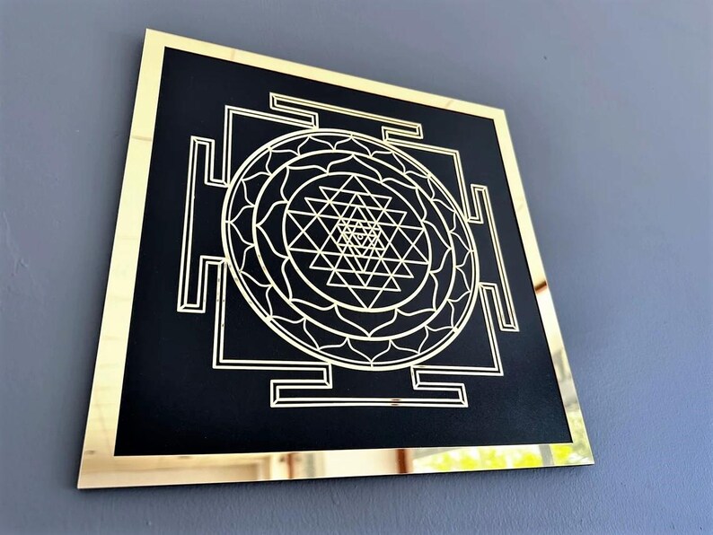 Puede incluir: Una pieza de arte de pared cuadrada con un marco dorado, fondo negro y un dise&ntilde;o geom&eacute;trico blanco. El dise&ntilde;o presenta un mandala central con un s&iacute;mbolo Sri Yantra.