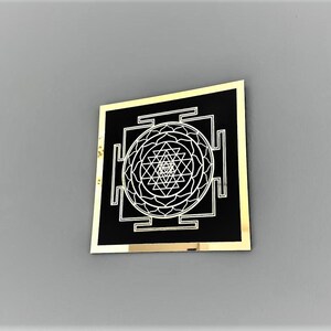 Puede incluir: Un cuadrado negro con un borde dorado, que presenta un dise&ntilde;o blanco de Sri Yantra. El Sri Yantra es un patr&oacute;n geom&eacute;trico complejo con un punto central y m&uacute;ltiples tri&aacute;ngulos.