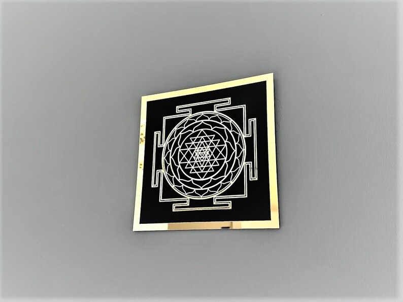 Puede incluir: Un cuadrado negro con un borde dorado, que presenta un dise&ntilde;o blanco de Sri Yantra. El Sri Yantra es un patr&oacute;n geom&eacute;trico complejo con un punto central y m&uacute;ltiples tri&aacute;ngulos.